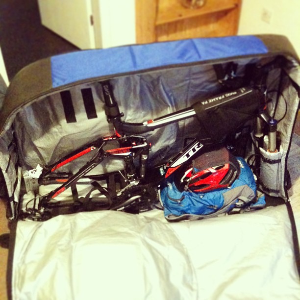 Partially disassembled bicycle packed in a large soft case with a helmet and blue bag beside it.