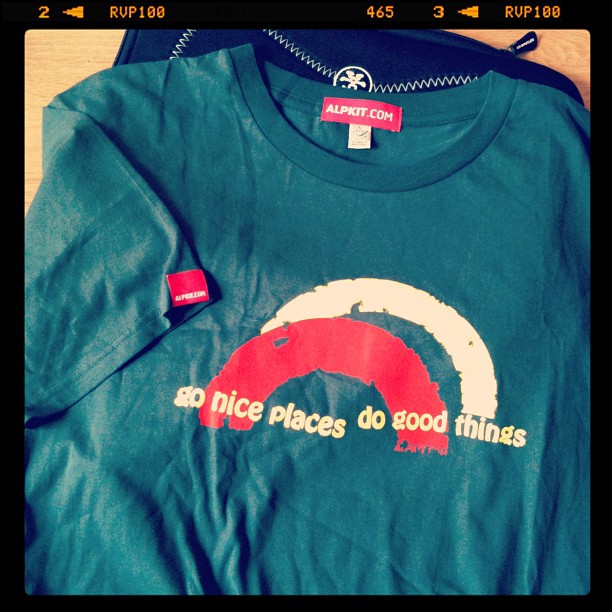 Blue ALPKIT T-shirt with a red and white rainbow graphic and the text “go nice places do good things,” laid on a dark bag.