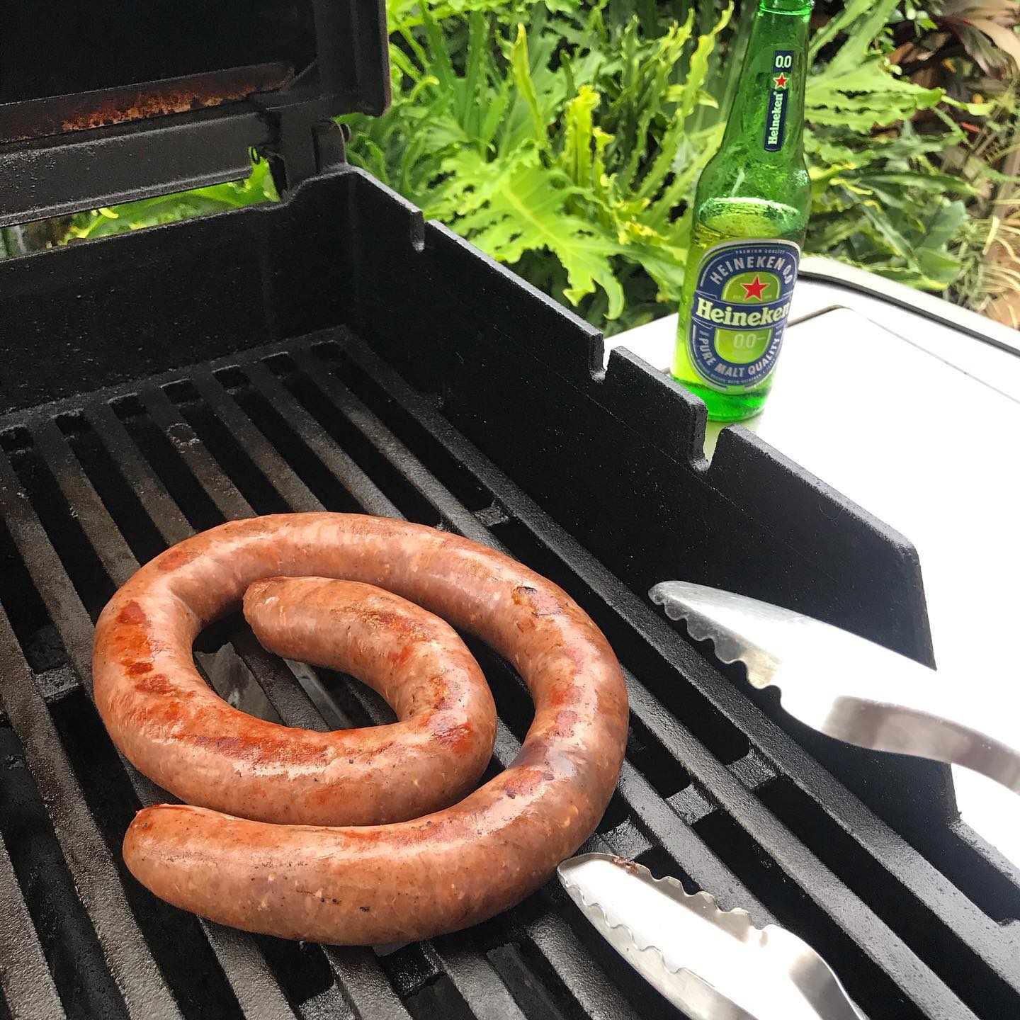 Coiled sausage browning on a grill with metal tongs beside it and a Heineken 0.0 bottle on a table against leafy greenery.