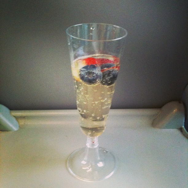 Clear plastic flute of sparkling drink with blueberries and a strawberry slice on a tray table against a gray seatback.