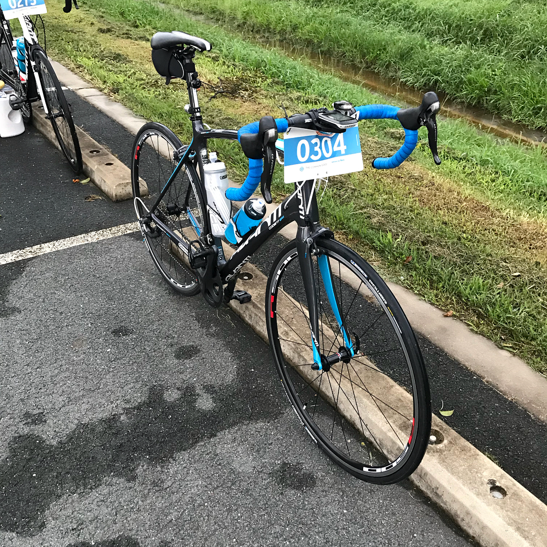 Black road bike with blue handlebar tape and race number 0304 parked beside a curb, with grass and a drainage ditch behind it.