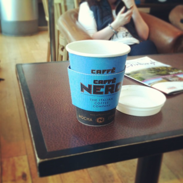 Blue Caffè Nero takeaway coffee cup on a cafe table with a white lid beside it and a seated person blurred in the background.