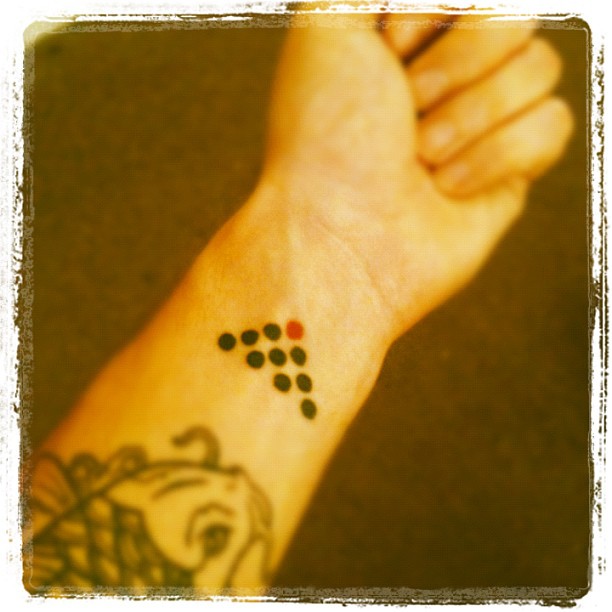 Inner wrist with a small dotted arrow tattoo in black and one red dot, plus part of a larger tattoo near the forearm.