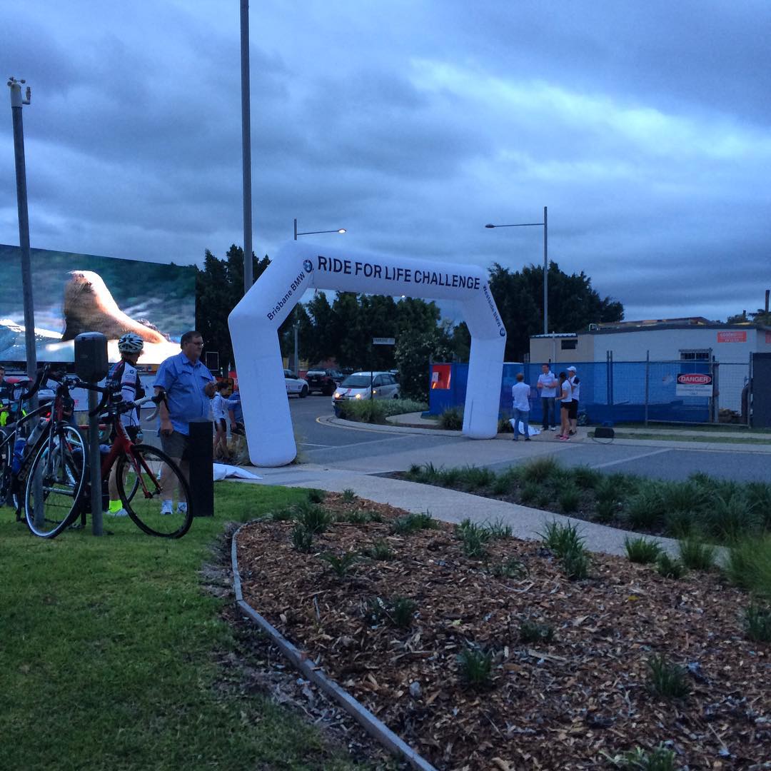 Inflatable "Ride for Life Challenge" arch by a roadway, with bicycles, a few people, and a large screen under a cloudy evening sky.