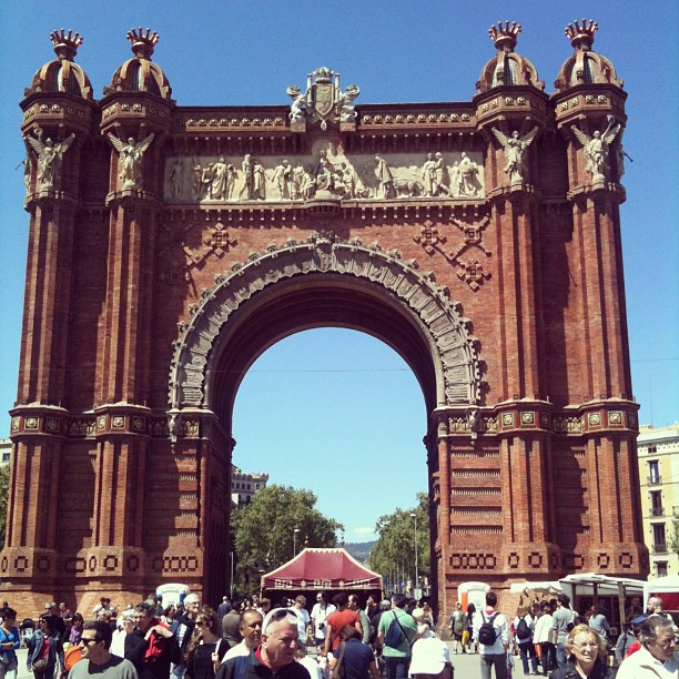 Large red brick triumphal arch with ornate sculptures and columns, framed by a blue sky, with a crowd gathered below.