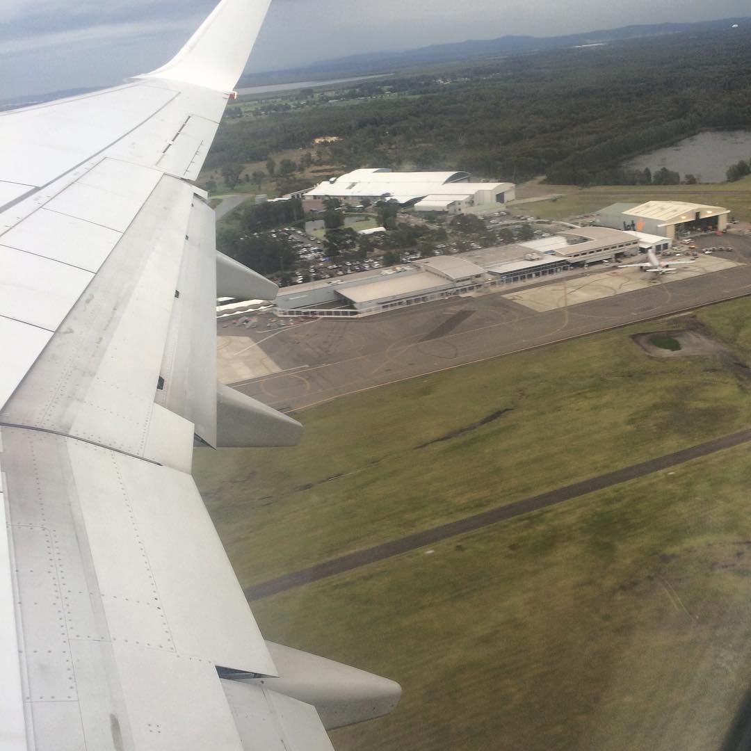 View from an airplane window showing the wing above an airport runway, terminal buildings, and surrounding grassy fields.