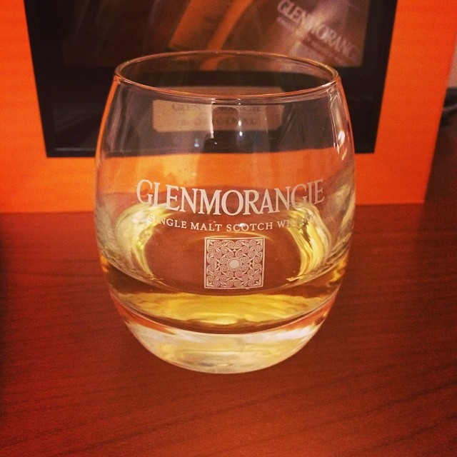 Glenmorangie whisky glass with a small pour of amber liquid on a wooden table, with an orange and black background.