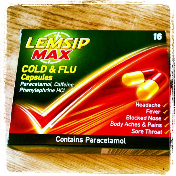 Box of Lemsip Max Cold & Flu Capsules on a wooden surface, showing red and yellow capsules and symptom relief text.