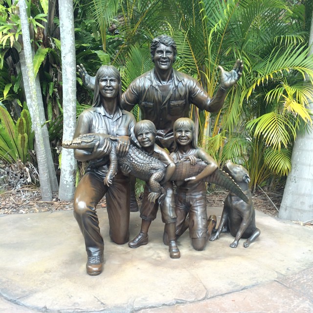 Bronze statue of two adults and two children posing with a crocodile, with a dog beside them and tropical plants behind.
