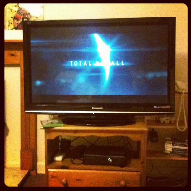Flat-screen TV on a wooden stand displaying the Total Recall title screen, with cables, a game console, and DVDs below.