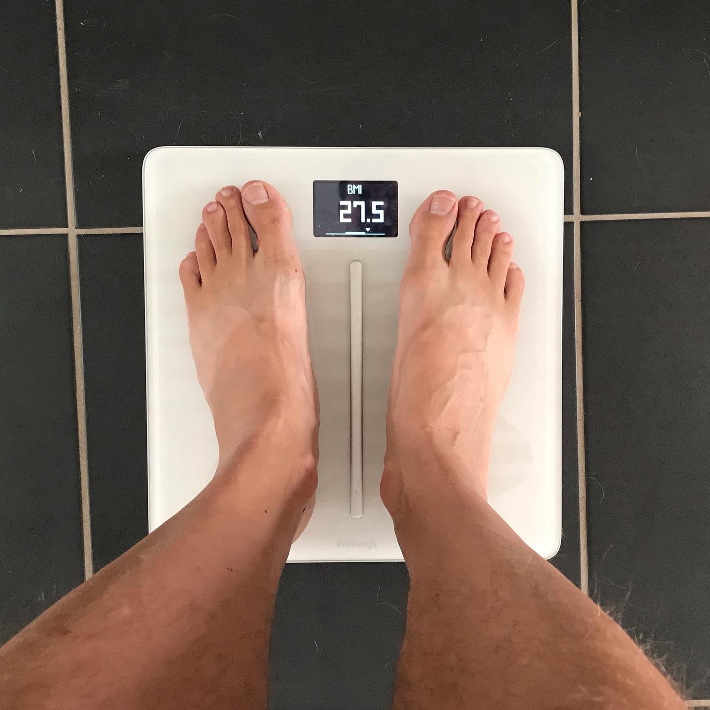 Bare feet standing on a white digital scale on black tile, with the display showing BMI 27.5.