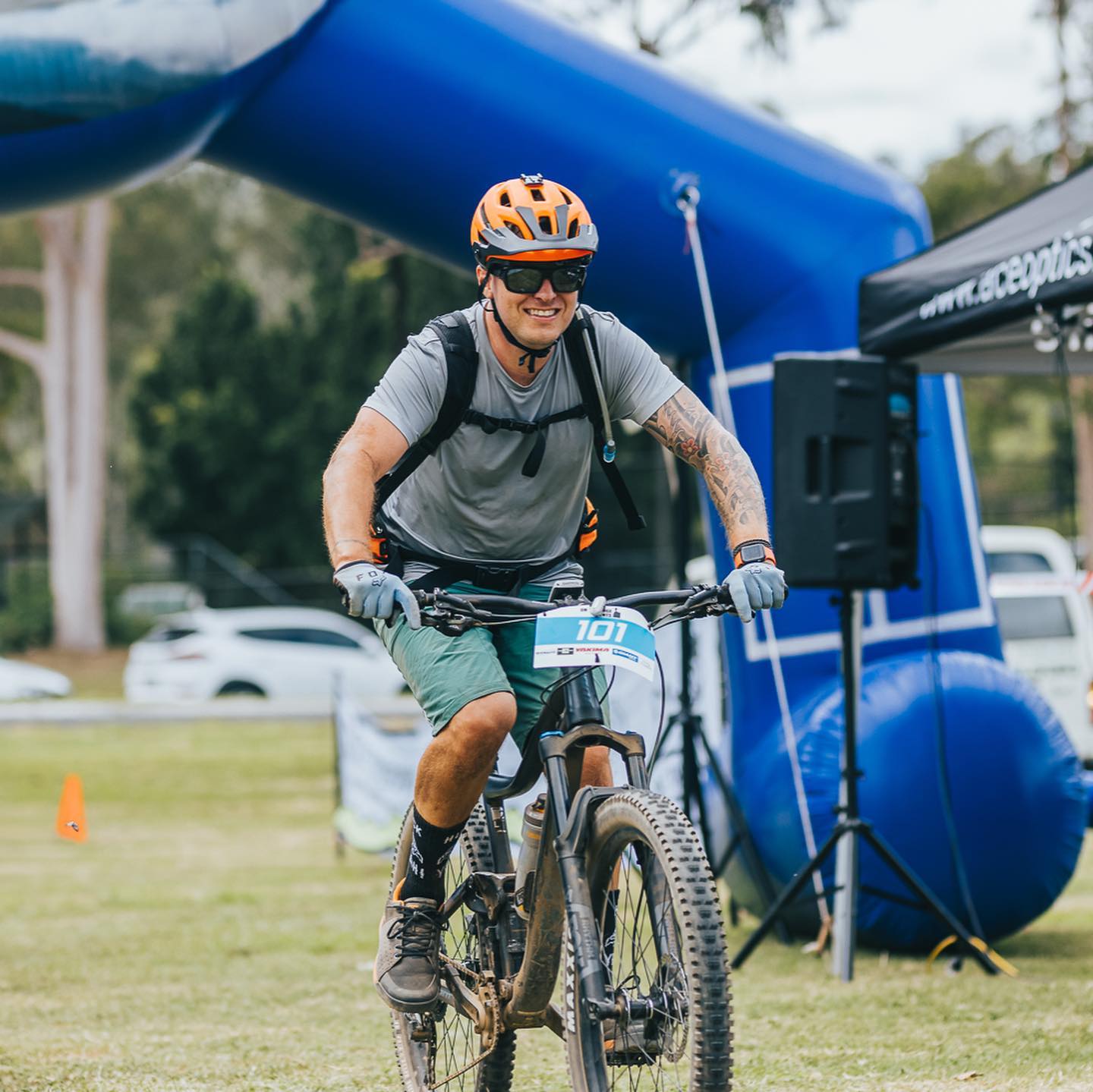 Smiling cyclist in an orange helmet rides a mountain bike on grass beneath a large blue inflatable arch.