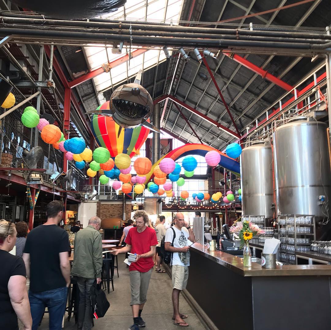 People walk through a large industrial-style hall with colorful hanging lanterns, a shark decoration, and stainless steel tanks beside a bar.