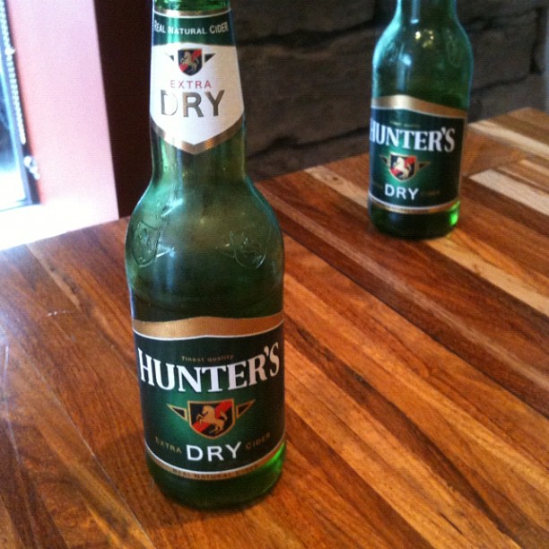 Two green Hunter’s Dry cider bottles on a wooden table, with one bottle in the foreground and one farther back.