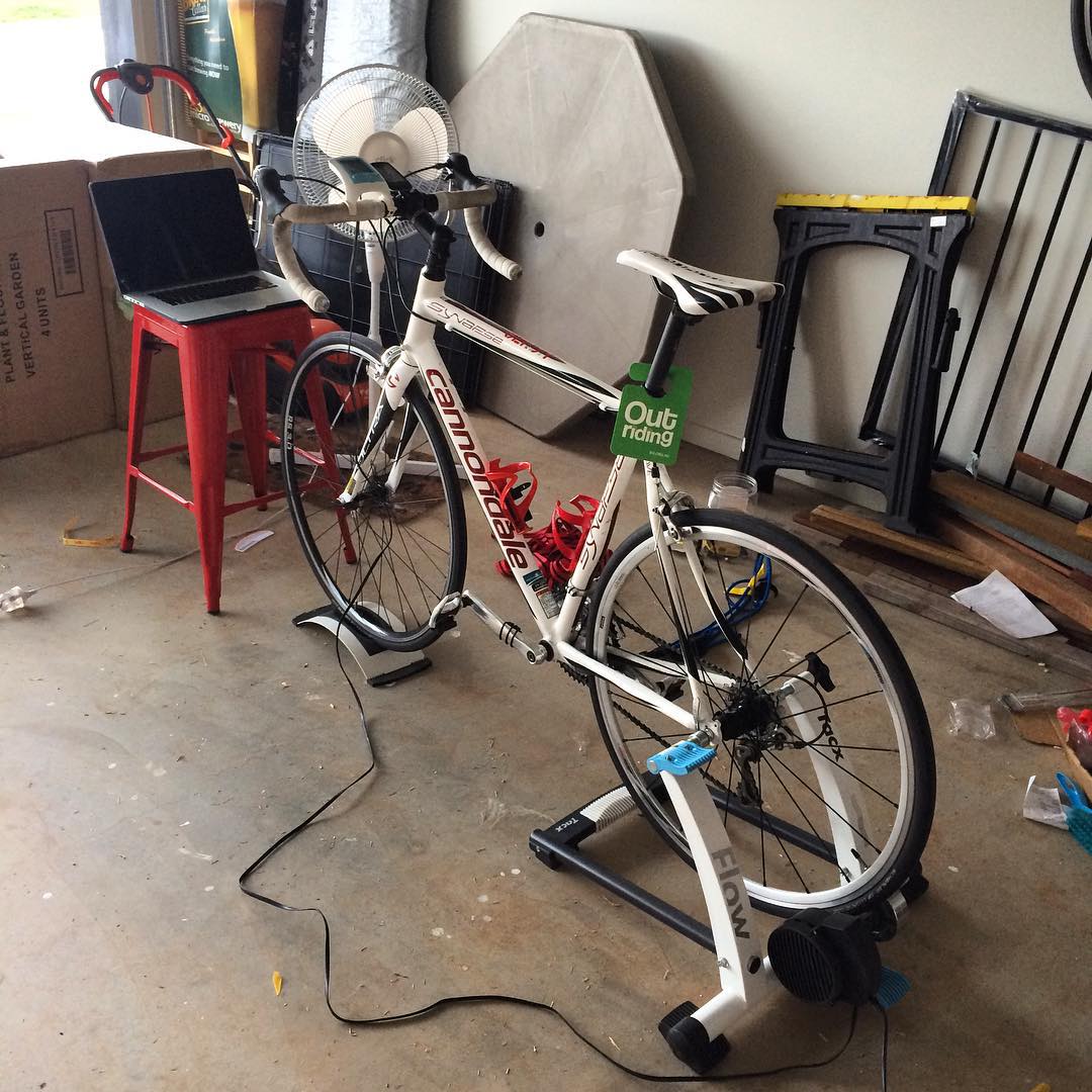 White road bike mounted on an indoor trainer in a garage, with a laptop on a red stool and a fan positioned in front.