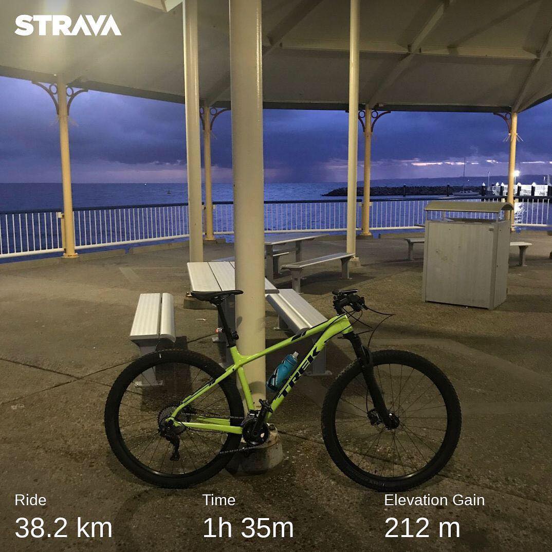 Bright green mountain bike under a covered seaside pavilion at dusk, with benches, ocean view, and Strava ride stats overlaid.