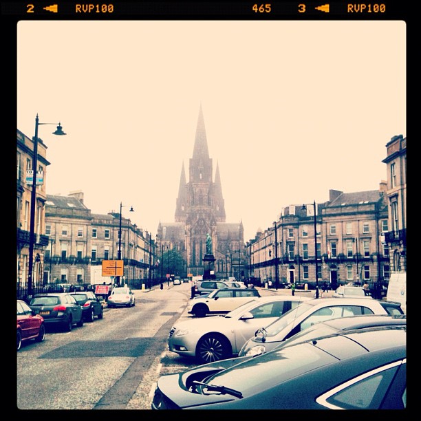 Foggy city street lined with parked cars and stone buildings, leading to a tall cathedral with a spire in the distance.