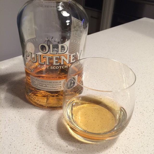 Partially full Old Pulteney whisky bottle beside a glass with a small pour of amber liquor on a light countertop.