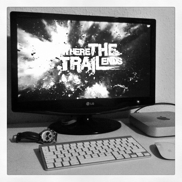 Black-and-white desk setup with an LG monitor displaying “HERE THE TRAIL ENDS,” a keyboard, mouse, watch, and a small desktop computer.