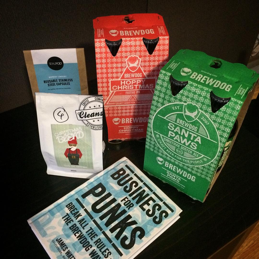 BrewDog holiday beer packs, a coffee bag, reusable stainless steel capsules, and a book arranged on a dark table.