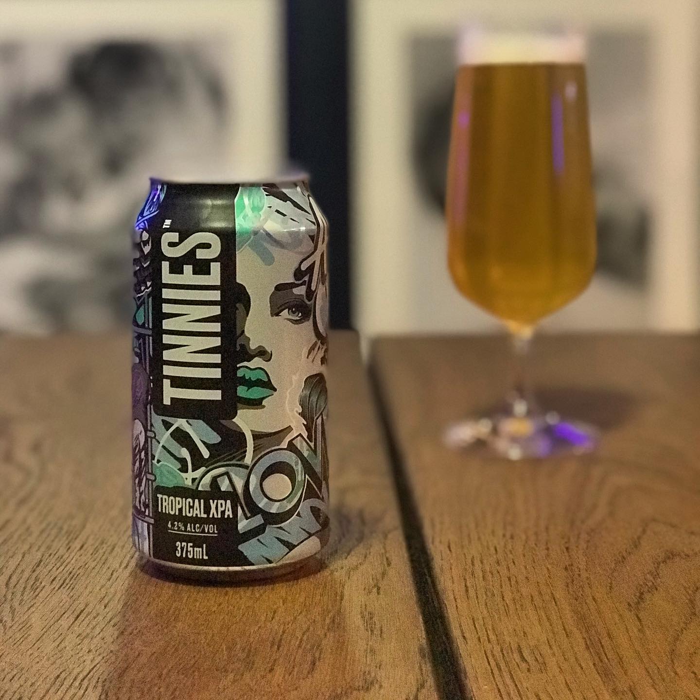 Can of Tinnies Tropical XPA on a wooden table with a filled beer glass blurred in the background.