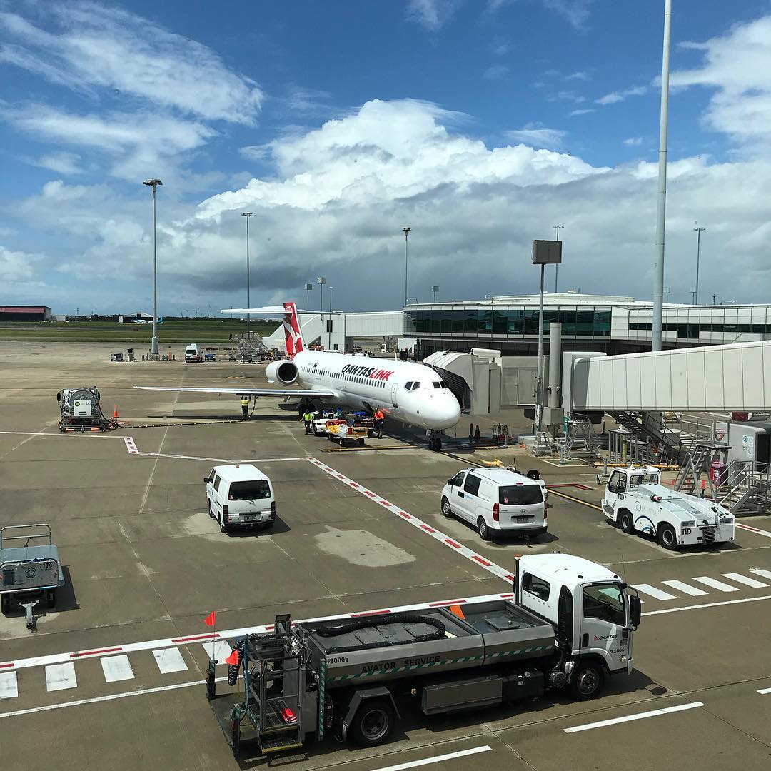 QantasLink jet parked at an airport gate with service vehicles on the tarmac under large white clouds and a blue sky.
