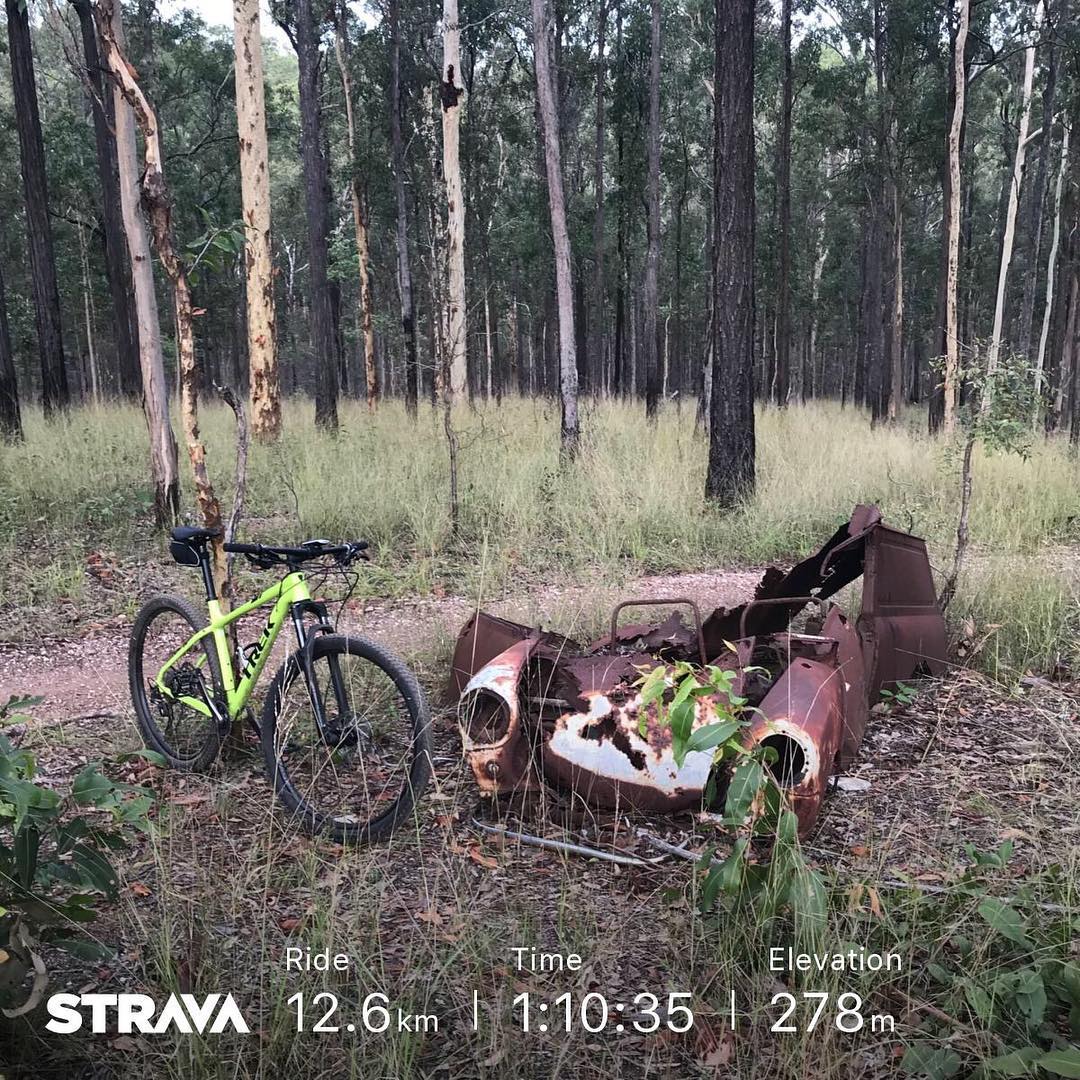 Bright green mountain bike beside a rusted stripped car body in a grassy forest, with Strava stats along the bottom.