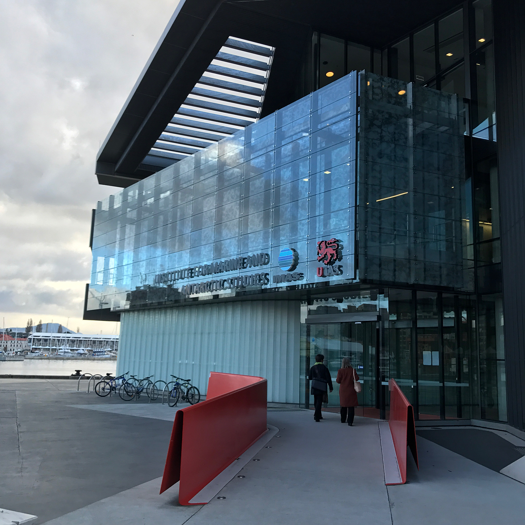 Modern glass building entrance with two people walking inside, red railings, bicycles, and cloudy sky by the waterfront.
