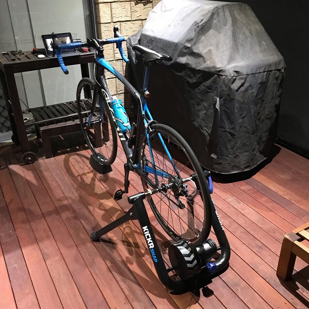 Blue road bike mounted on an indoor trainer on a wooden deck beside a covered grill and small table.