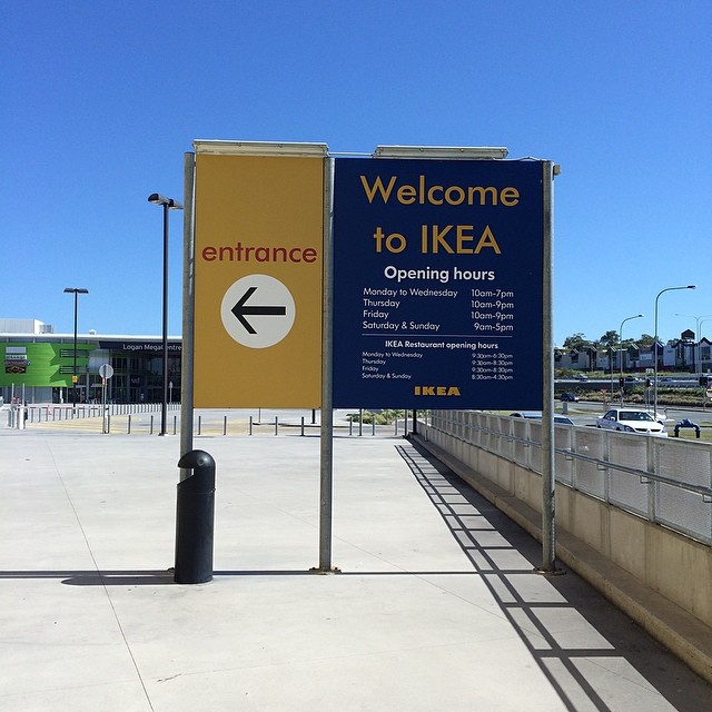 IKEA entrance sign with a left arrow and opening hours on a sidewalk under a clear blue sky.