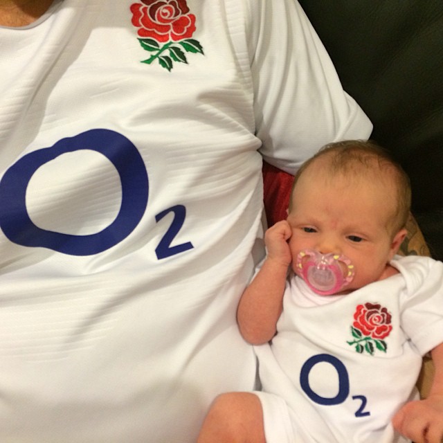Baby with a pacifier beside an adult, both wearing white shirts with a red rose and blue O2 logo.