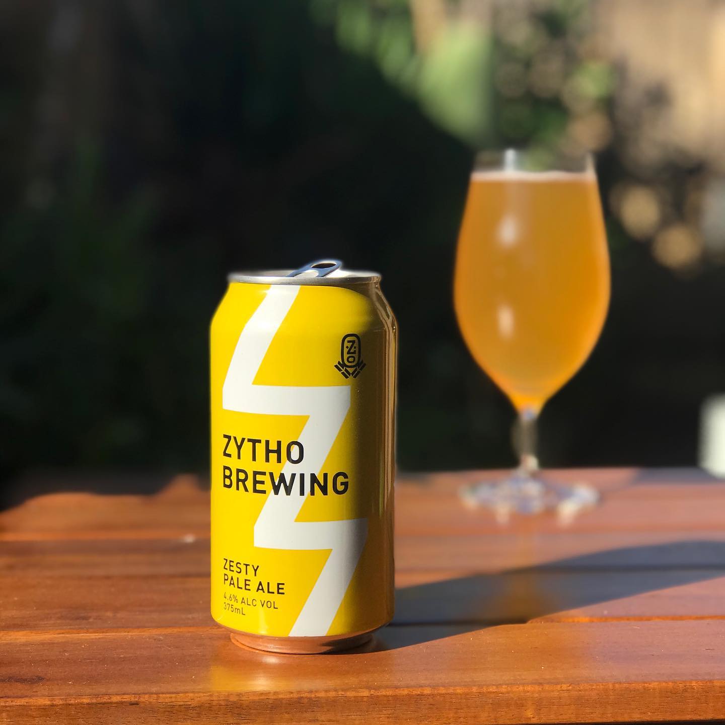 Yellow Zytho Brewing zesty pale ale can on a wooden table, with a filled stemmed glass blurred in the background.