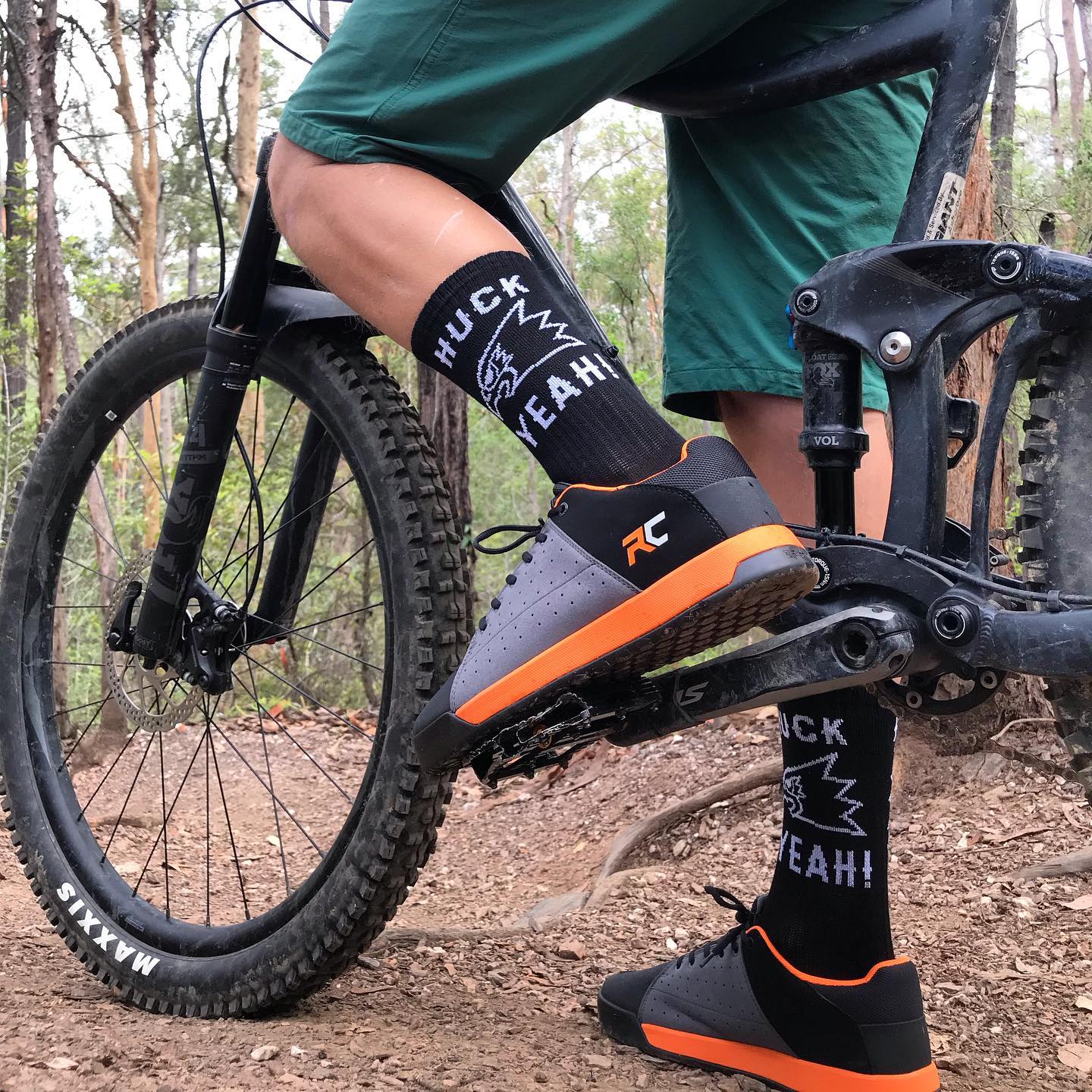 Close-up of a mountain biker’s legs, black “Huck Yeah!” socks, and black-and-orange shoes on a trail in the woods.