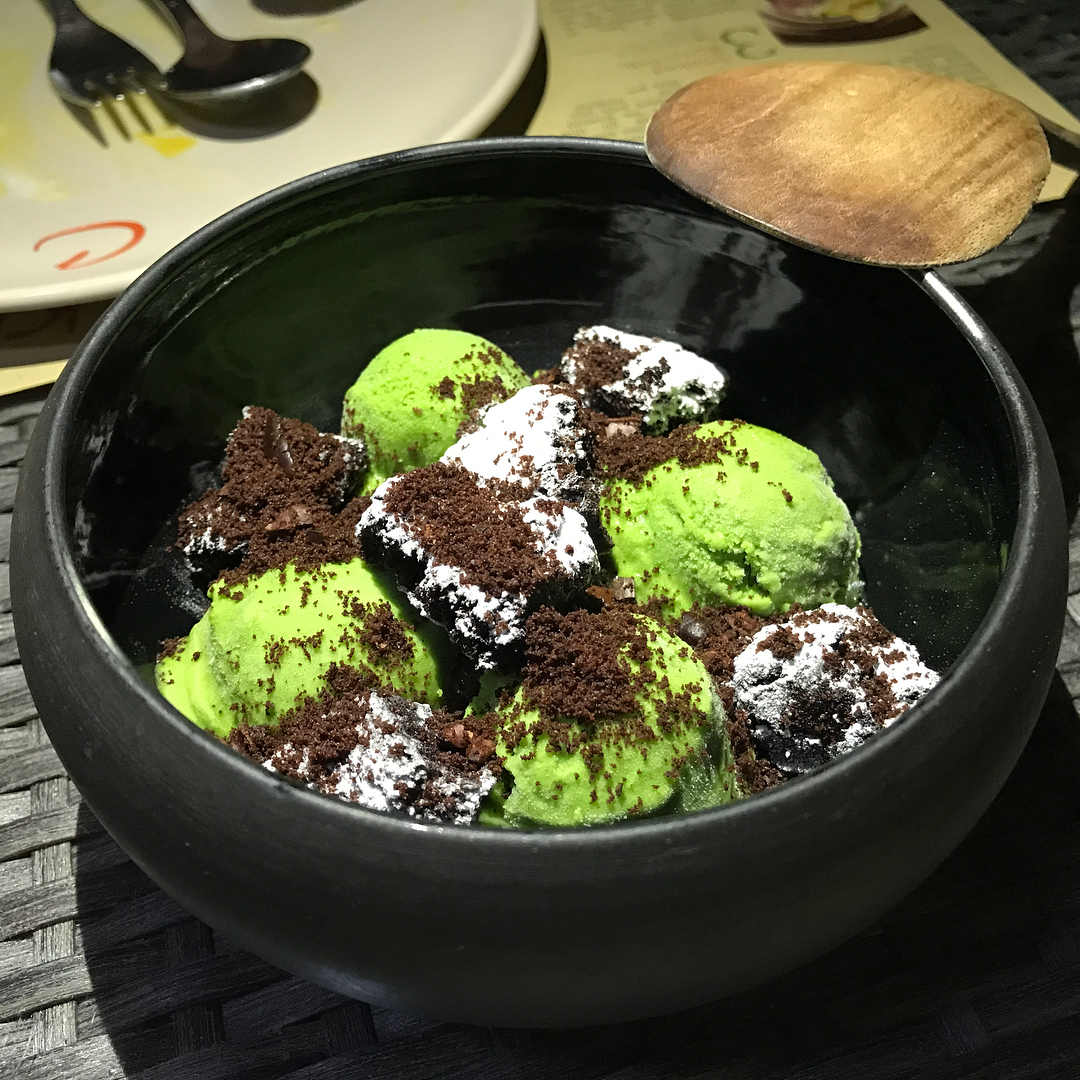 Black bowl of bright green ice cream scoops topped with chocolate crumbs and powdered sugar, with a wooden spoon resting on the rim.