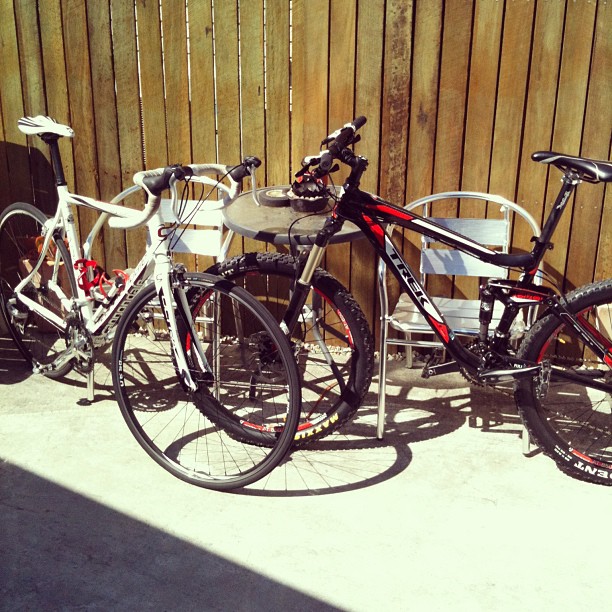 White road bike and black mountain bike parked beside a small metal table and chairs against a wooden fence.