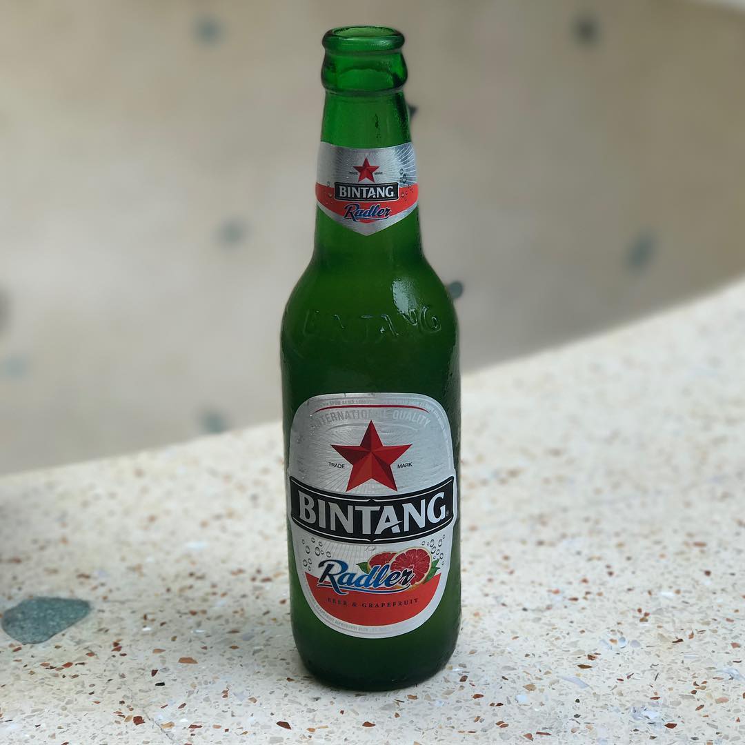 Green Bintang Radler beer and grapefruit bottle standing on a speckled surface against a blurred light background.