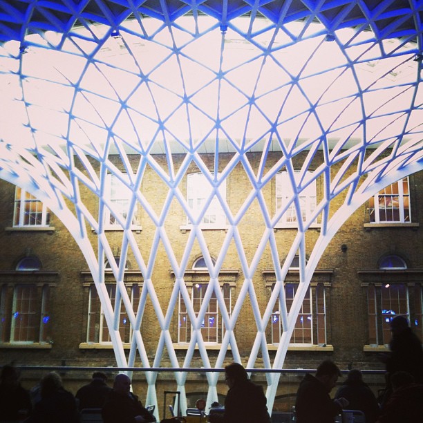 White lattice canopy over seated silhouettes in an indoor courtyard, with a brick building facade and windows in the background.