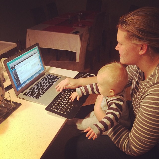 Woman holding a baby while using a laptop and separate keyboard at a table.