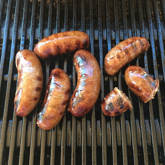 Six browned sausages grilling on black metal grates, with one sausage cut into two pieces.