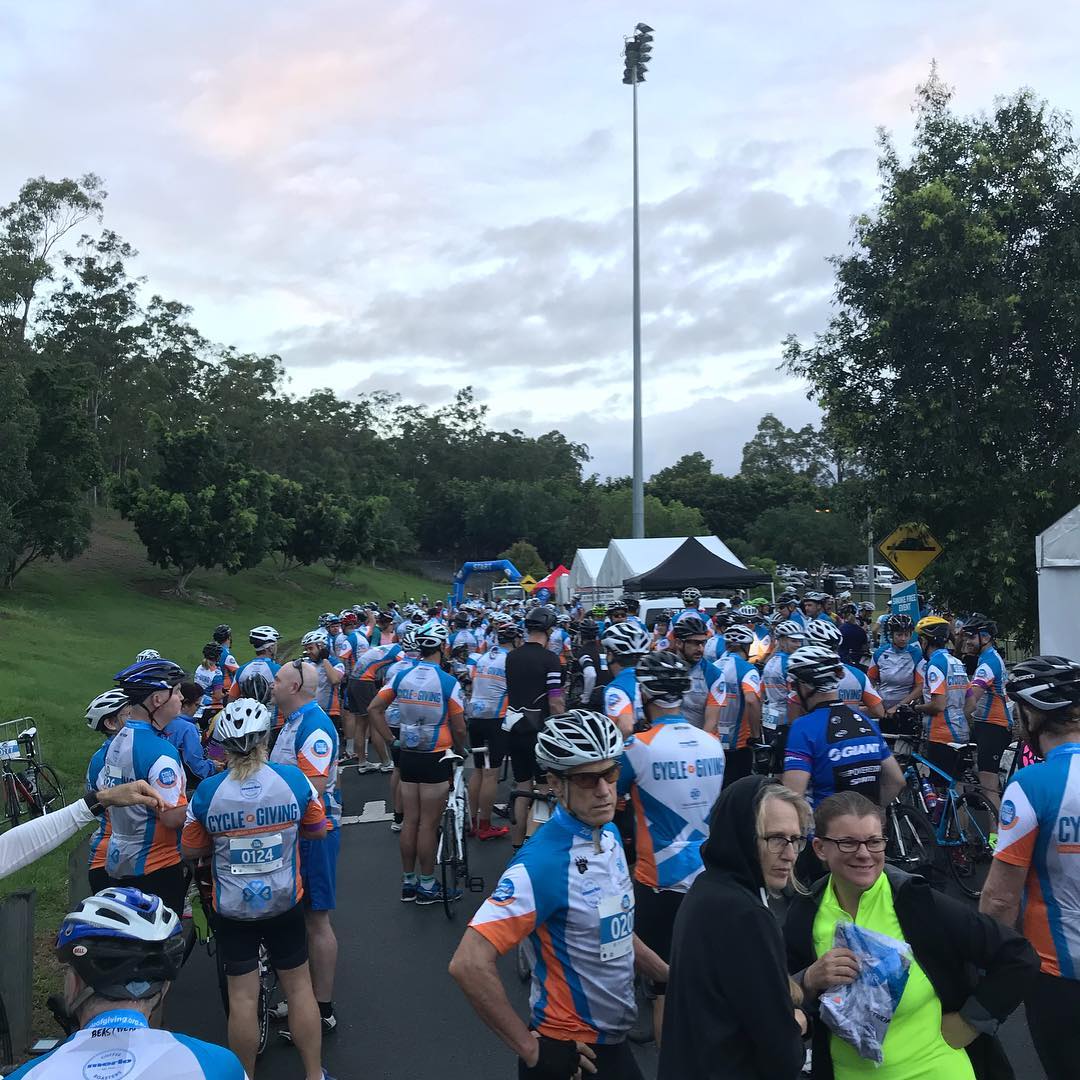 Crowd of cyclists in matching jerseys and helmets gather on a road near tents and trees under a cloudy sky.