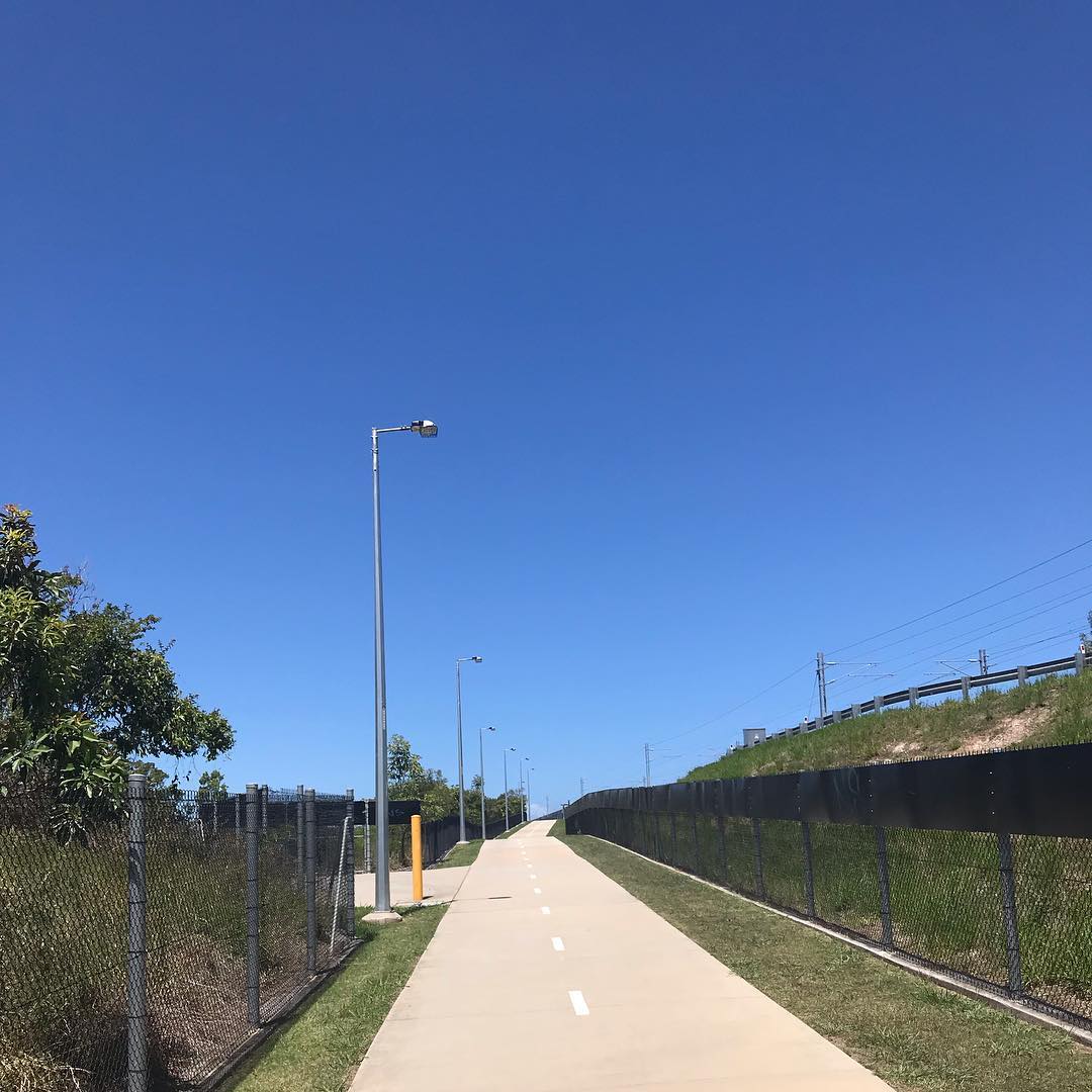 Paved path with dashed center line between chain-link fences, lined with streetlights under a clear blue sky.