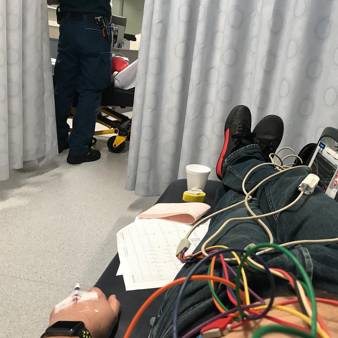 Person reclines on a hospital bed with ECG wires and an IV in hand while a staff member stands behind a curtain nearby.