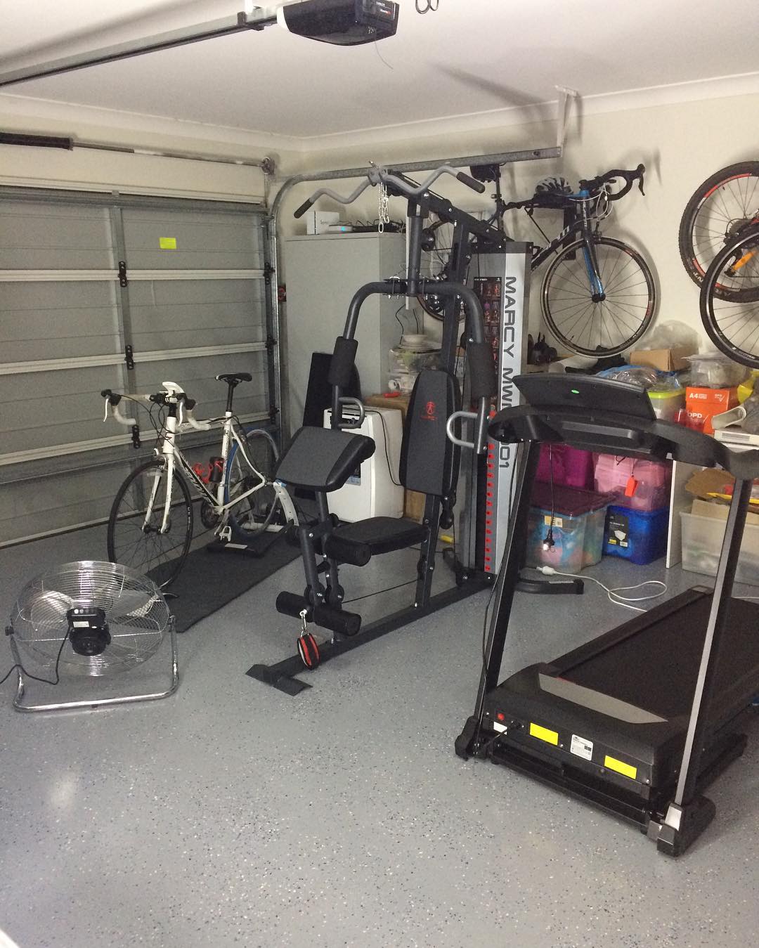 Garage gym with a treadmill, weight machine, floor fan, white road bike, and two bicycles mounted on the wall.