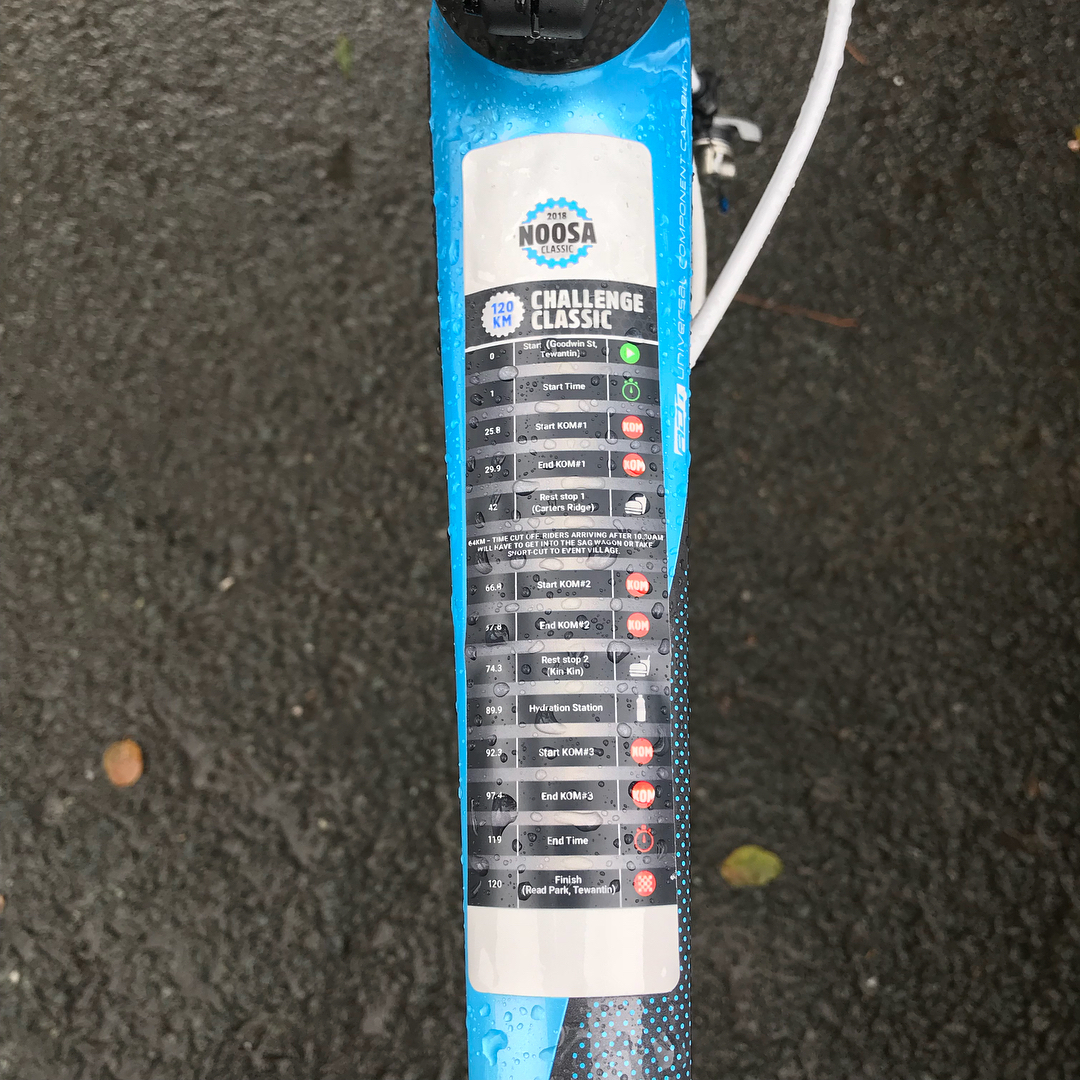 Close-up of a blue bicycle top tube with a Noosa Challenge Classic route sticker covered in raindrops on wet pavement.