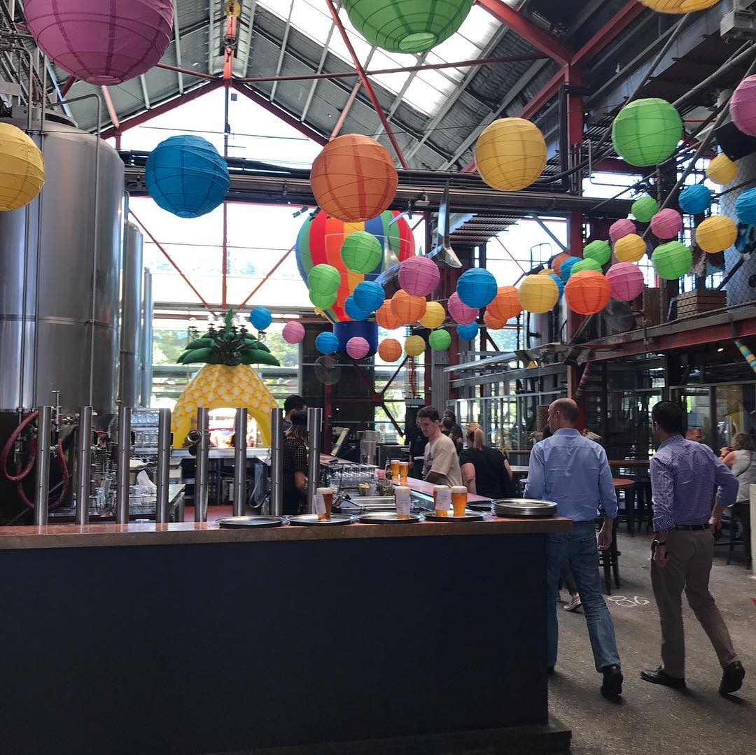 Industrial-style bar interior with colorful hanging lanterns, a pineapple arch, stainless tanks, and people standing near the counter.