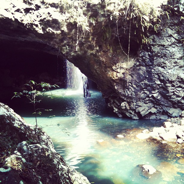Small waterfall inside a rocky cave opening, flowing into a turquoise pool with sunlight reflecting on the water.