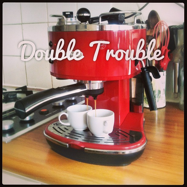 Red espresso machine pouring coffee into two small white cups, with “Double Trouble” written across the front.