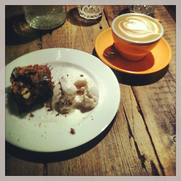 Slice of cake and a scoop of melting ice cream on a white plate beside a latte in an orange cup on a wooden table.