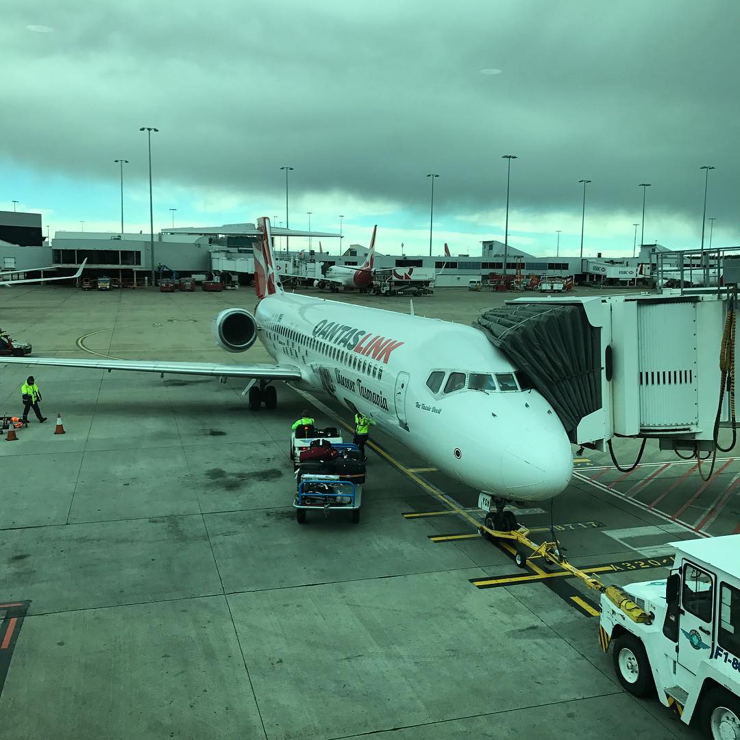QantasLink airplane at an airport gate with a jet bridge attached, luggage carts nearby, and dark clouds overhead.