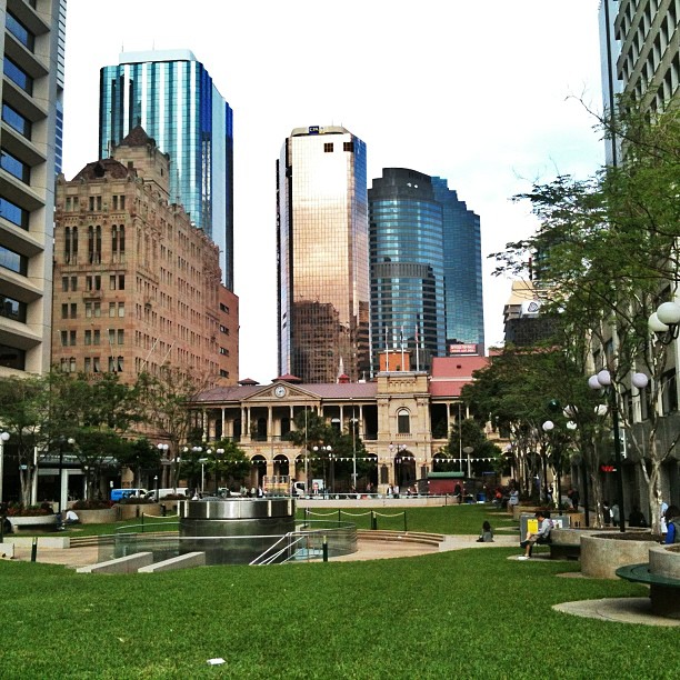 Urban park with green lawn and trees in front of historic and modern high-rise buildings.